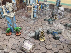 A top down shot of a Battletech map. Visible mechs on the opposing side along the top of the map: Hunchback (partially obscured by buildings), Bushwacker (also partially obscured by building), Berserker (head and shoulders cropped off), Nightsky with Commando hiding behind it. My forces from left to right along the middle: Assassin and Spartan hiding out for an ambush, Charger and Ostscout capturing objectives.