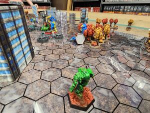 A close up photo of a Battletech map, showing an Assassin (blurry, foreground) firing on a now-dead Commando. In the distance, many mechs committing violence against my beautiful Spartan.