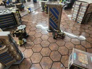A close up photo of a Battletech map. My Ostsol lies dead and destroyed next to a building. My Charger has activated a point for scoring. In the distance, a Crusader and Hunchback look on, while my Hierofalcon takes a shot at the rear armour of the Hunchback.